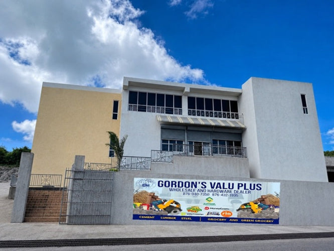 A modern two-story white building with a beige side wing, featuring a large outdoor banner for 'Gordon's Valu Plus' on the lower wall in front, set against a bright blue sky with white clouds.